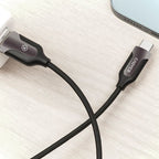 A black braided USB-C to USB-C charging cable with a gray and black connector housing, shown plugged into a white charger on a light wood surface next to a smartphone.