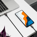 A black USB-C cable with a visible 'SAMSUNG' logo on the connector, shown plugged into a smartphone and a laptop on a white desk. The smartphone screen displays a colorful abstract pattern.