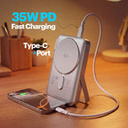 A silver 35W PD fast-charging wireless power bank with a built-in stand, shown on a wooden desk with a smartphone charging in its Type-C port. The image features promotional text overlays highlighting its features.