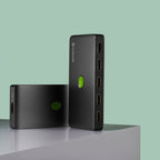 Two black CARVCES electronic adapters, one standing upright and one lying down, featuring a green circular indicator light and multiple HDMI ports, set against a solid sage green background.