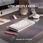 A white low-profile wireless keyboard shown on a dark desk with a mouse, computer monitor, mug, and pen. White text overlays read 'LOW-PROFILE KEYS' and 'MINI SIZE.