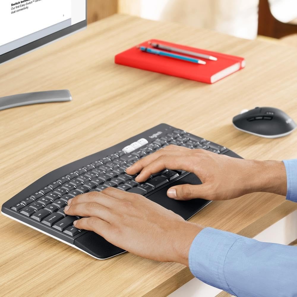 Person using a wireless keyboard on a desk with a computer monitor, notebook, and mouse.