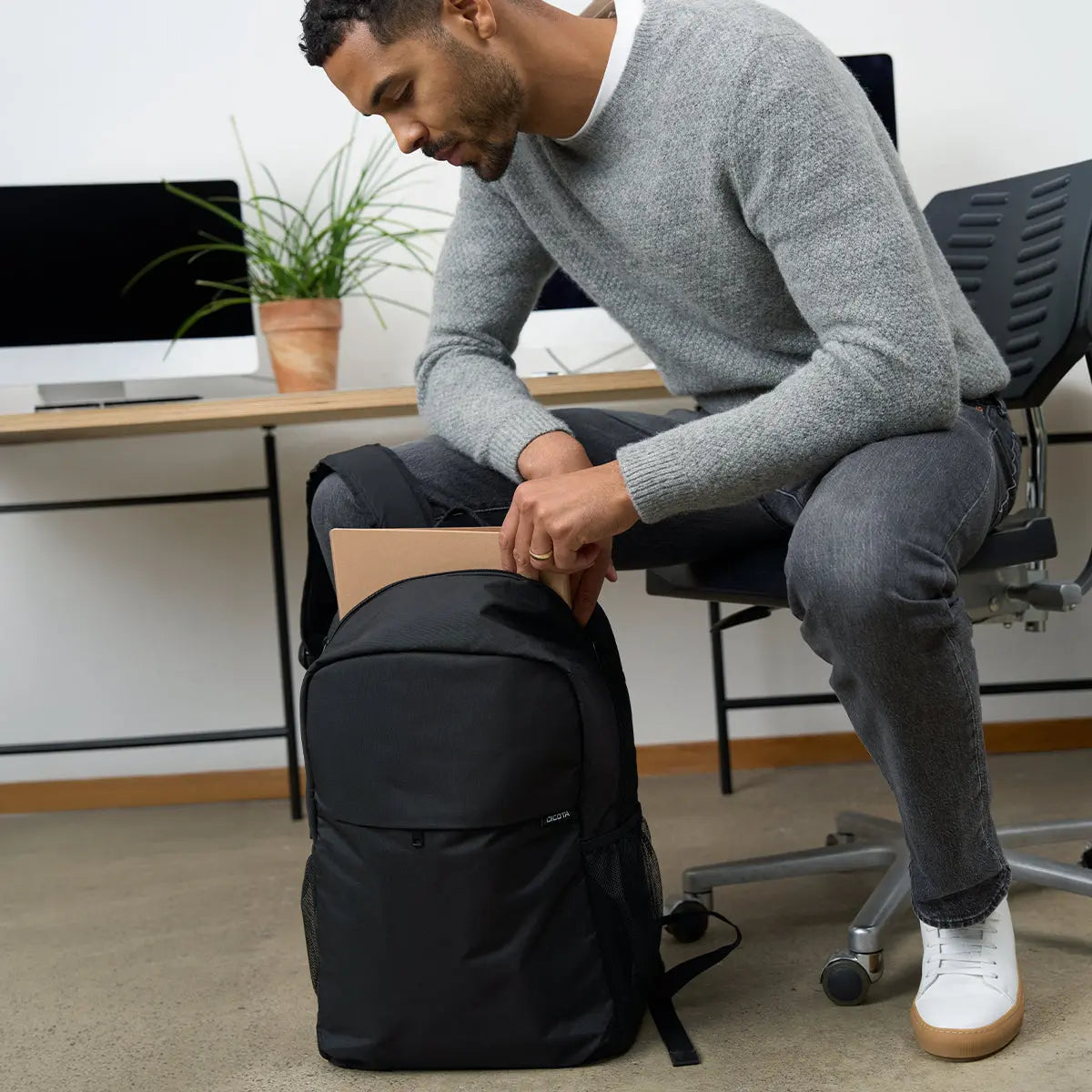 A man in a gray sweater and jeans sits on an office chair, placing a brown folder into a black backpack on the floor in a modern office setting.