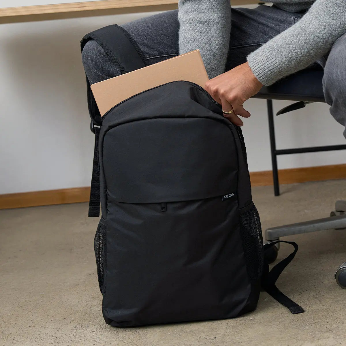 A black backpack with a zippered top pocket and side pockets, shown on the floor next to a person sitting on a chair and placing a tan folder into the top compartment.