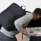 A man wearing a gray knit sweater and jeans carries a black backpack on his back while leaning over a desk in front of a computer monitor.