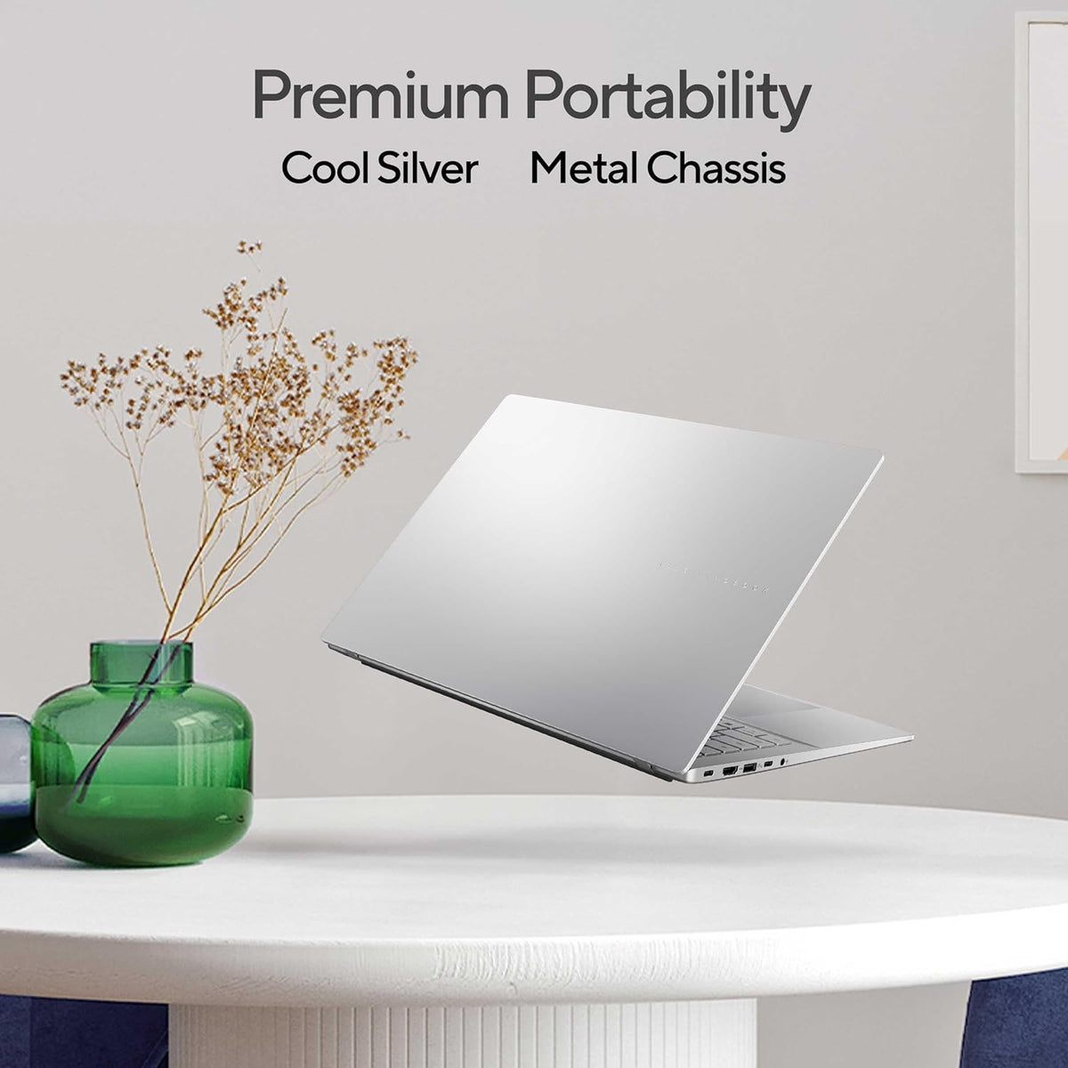 A silver laptop floating above a white table with a green glass vase and dried stems, with text reading 'Premium Portability Cool Silver Metal Chassis' on a light gray wall.
