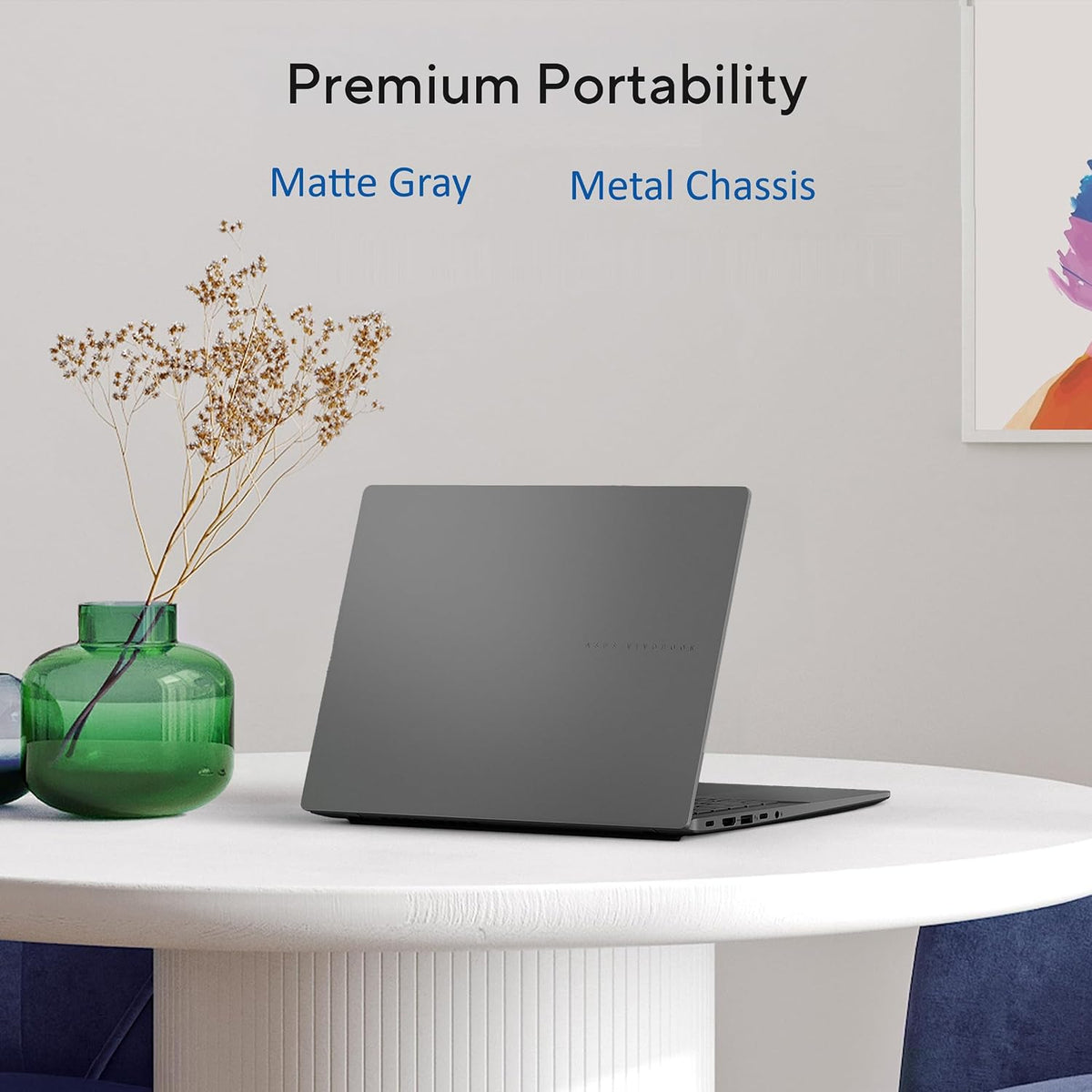 A matte gray laptop on a white table in a styled interior setting, with text reading 'Premium Portability' and 'Matte Gray Metal Chassis' above it. A green vase with dried stems sits to the left.