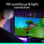 A Samsung computer monitor on a desk showing a video game scene, with a webcam mounted on top. A person is seated in front at an angle, viewed from behind, in a room with purple ambient lighting and acoustic panels on the wall.