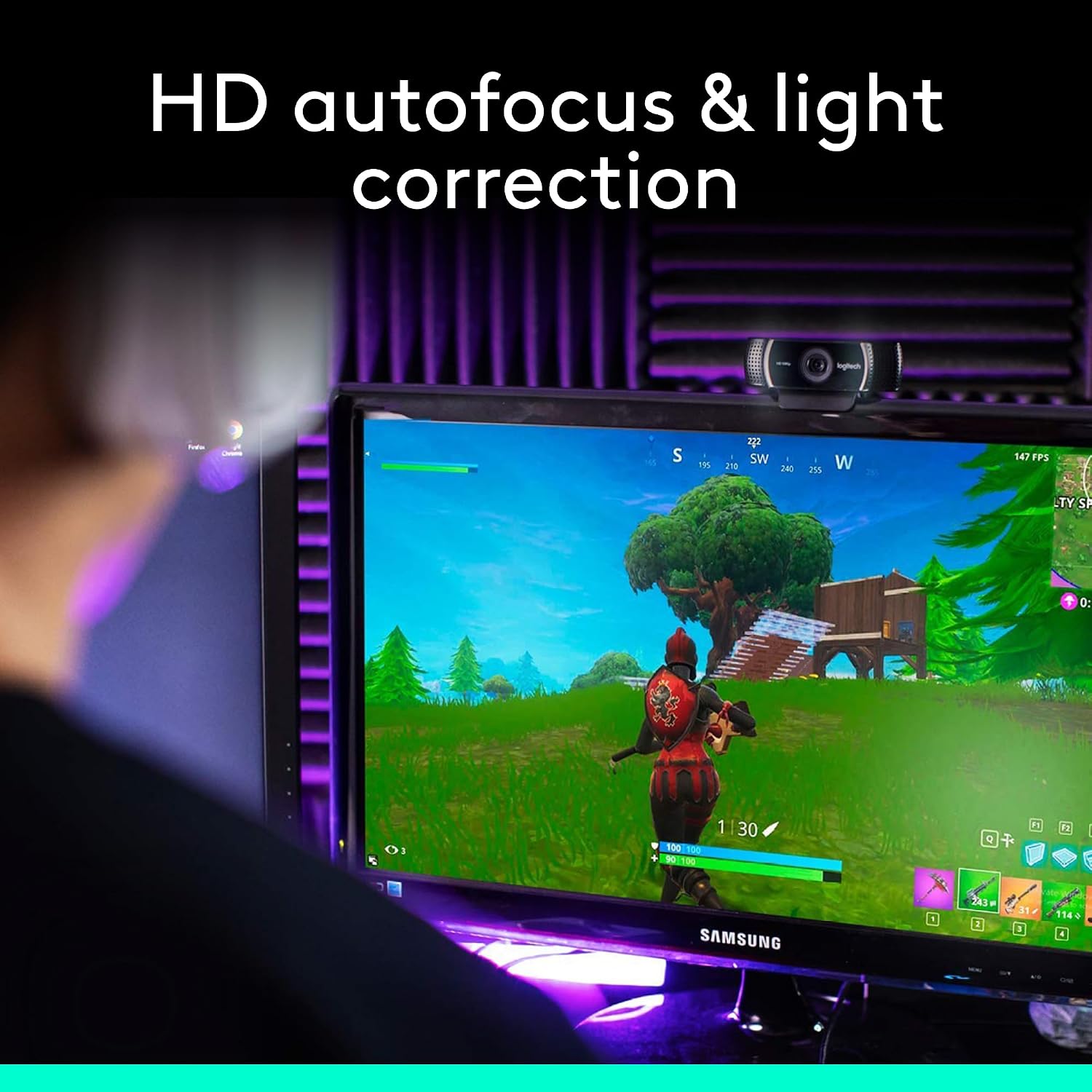 A Samsung computer monitor on a desk showing a video game scene, with a webcam mounted on top. A person is seated in front at an angle, viewed from behind, in a room with purple ambient lighting and acoustic panels on the wall.