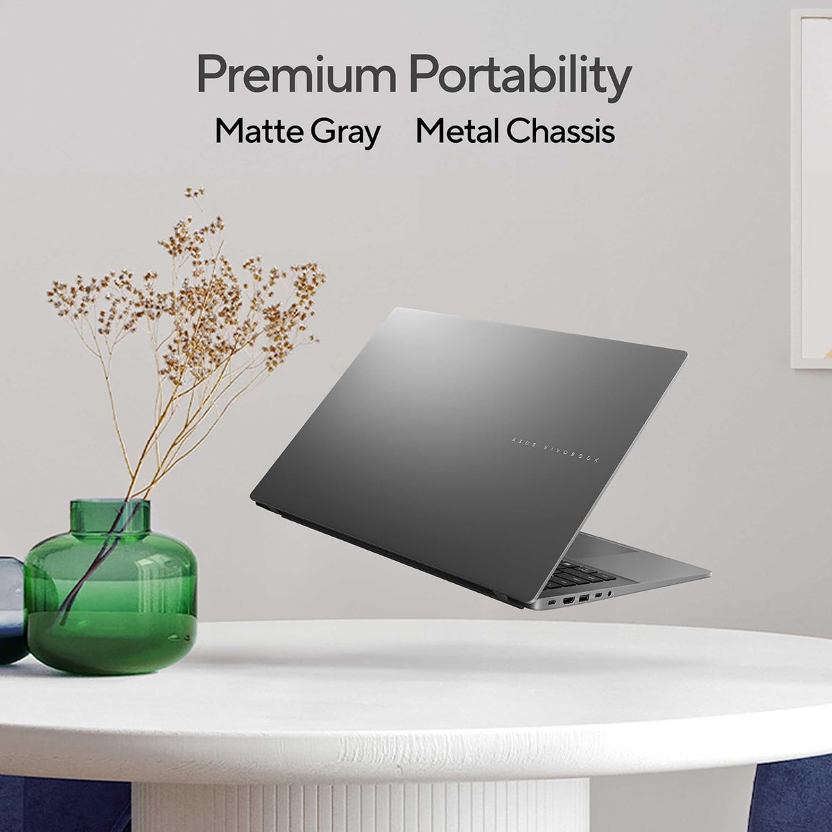 A matte gray metal laptop shown floating above a white table in a styled interior scene with dried flowers in a green vase and wall text reading 'Premium Portability Matte Gray Metal Chassis'.