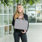 Woman holding a gray laptop bag in an indoor setting with large windows and plants.