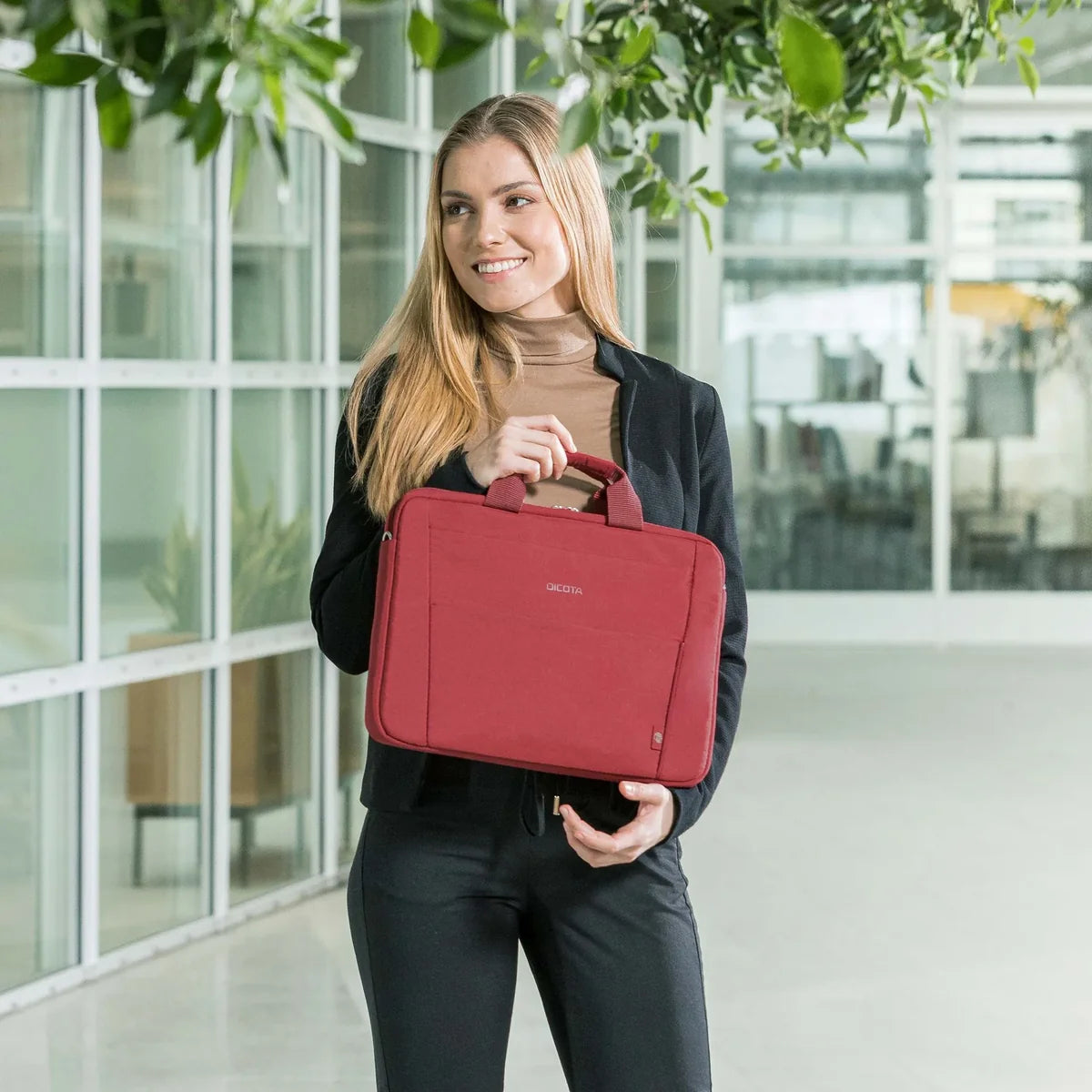 Woman holding a red bag in an indoor setting with large windows and plants.