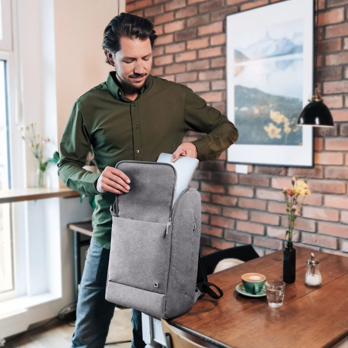 Man in a green shirt packing a gray backpack in a room with a brick wall and a window.