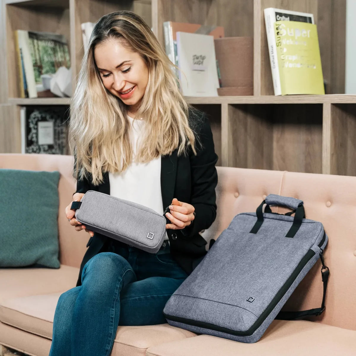A woman sits on a beige sofa holding a gray laptop sleeve, with a matching gray laptop bag placed beside her on the couch. The setting is a cozy living room with wooden bookshelves in the background.