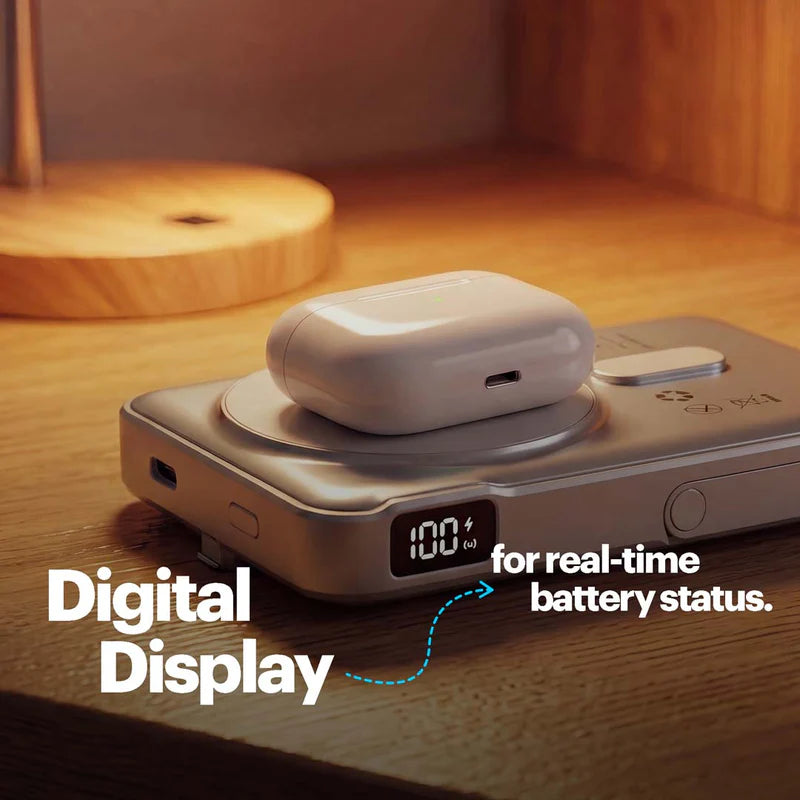 A silver electronic battery/power bank device sits on a wooden surface with a white wireless earbud case resting on top. The device has a small digital display showing 100% battery, and overlaid text reads 'Digital Display' and 'for real-time battery status.