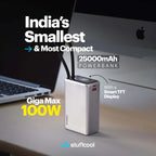 A silver power bank with a small display and two antennas, shown on a desk next to a laptop and computer monitor. Text overlays read 'India's Smallest & Most Compact', '25000mAh POWERBANK', 'Giga Max 100W', and 'With a Smart TFT Display'.