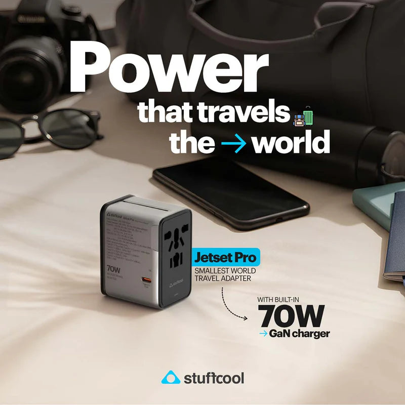 A Jetset Pro 70W GaN travel charger by stuffcool shown on a beige fabric surface with travel props like a phone, sunglasses, camera, and bag in the background. Large text reads 'Power that travels the world' and 'Jetset Pro Smallest World Travel Adapter with built-in 70W GaN charger.