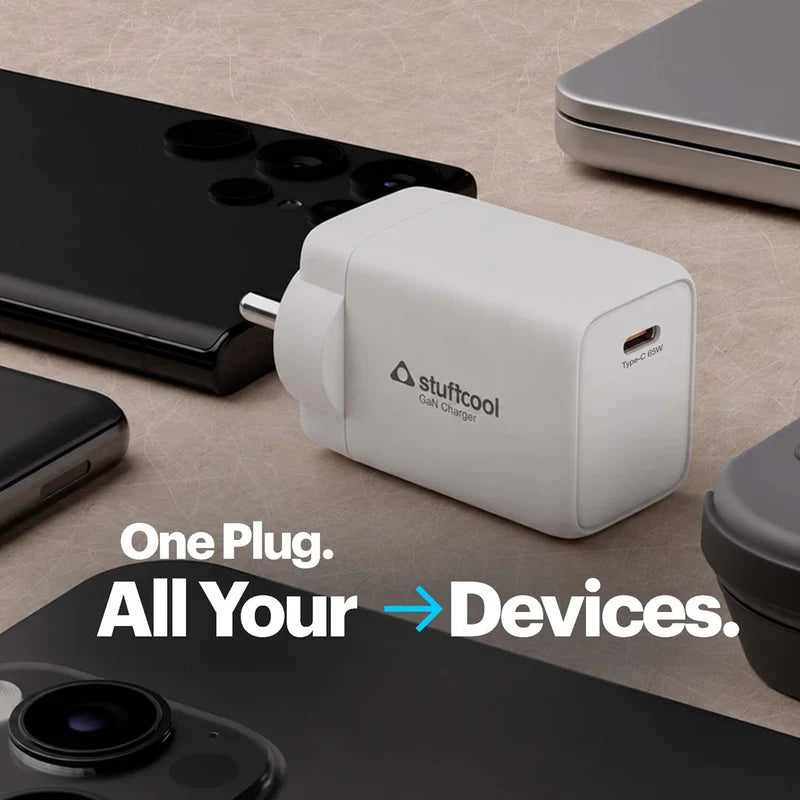A white Stuffcool USB charger plug shown on a beige surface with several smartphones and a laptop around it. Large text reads 'One Plug.