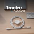 A white braided USB-C cable with a central hub connector, coiled on a light wood desk next to a laptop and keyboard. Large text overlay reads '1metre LONG CABLE'.
