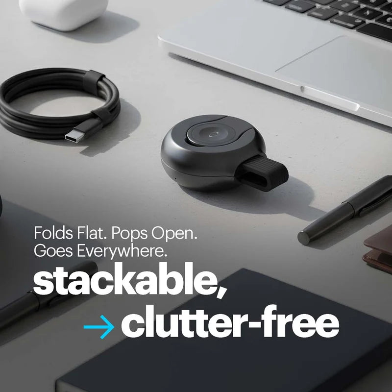 A small round black electronic accessory with a latch, shown on a desk beside a laptop, cable, pen, and notebook. Large overlaid text reads, 'Folds Flat.