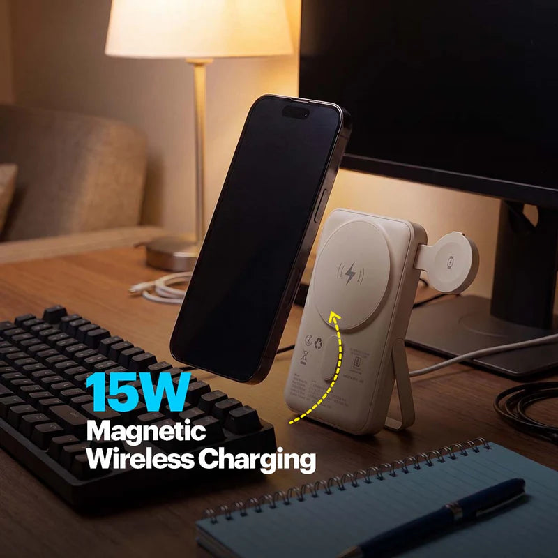 A white magnetic wireless phone charger standing on a wooden desk, charging a black smartphone. The scene includes a keyboard, lamp, monitor, and notebook in a home office setting, with text overlay reading '15W Magnetic Wireless Charging'.