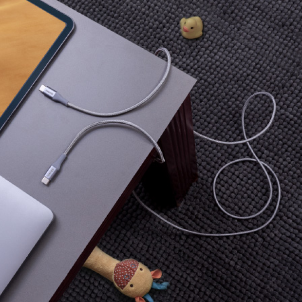 A gray braided USB-C charging cable draped across a dark gray desk surface next to a laptop, with a textured dark carpet in the background and small decorative objects nearby.