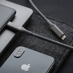 A close-up of a gray fabric charging cable with a metal connector labeled CADYCE, shown plugged into a dark gray laptop and phone on a dark textured surface.