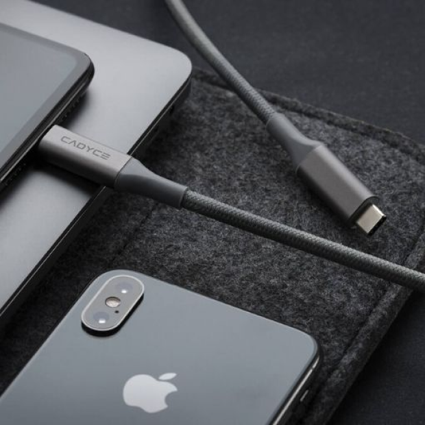 A close-up of a gray fabric charging cable with a metal connector labeled CADYCE, shown plugged into a dark gray laptop and phone on a dark textured surface.