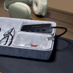 A black Logitech USB hub with multiple ports and a connected black cable, placed in a gray felt organizer tray on a desk. Glasses, keys, and a watch are visible in the tray, with headphones blurred in the background on a wooden surface.