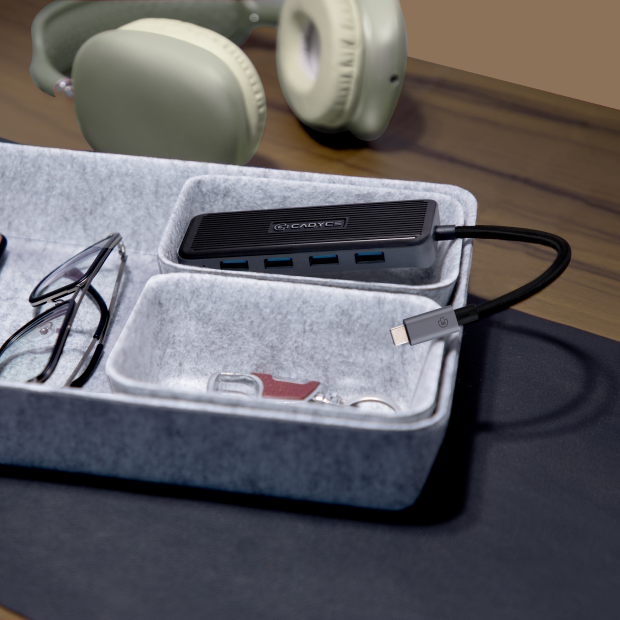 A black Logitech USB hub with multiple ports and a connected black cable, placed in a gray felt organizer tray on a desk. Glasses, keys, and a watch are visible in the tray, with headphones blurred in the background on a wooden surface.