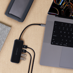 A black Gauronk multi-port USB charger adapter plugged into a gray laptop, shown in a styled flat-lay on a beige textured surface with a phone and notebook nearby.