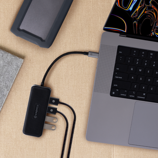 A black Gauronk multi-port USB charger adapter plugged into a gray laptop, shown in a styled flat-lay on a beige textured surface with a phone and notebook nearby.