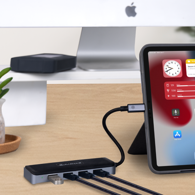 A black and silver USB-C hub with multiple ports sits on a light wood desk, connected to a tablet and other devices. The background shows a blurred iMac on a desk stand.