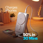 A silver Qi wireless power bank shown on a wooden desk, charging a smartphone. The power bank is displayed on a small stand, with a white cable plugged in.