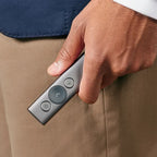 Hand holding a silver remote control with buttons, wearing beige pants and a blue jacket.