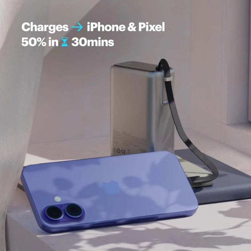 A portable rectangular power bank with a black wrist strap sits behind a blue smartphone on a light surface. Text in the upper left reads 'Charges -> iPhone & Pixel 50% in 30mins'.