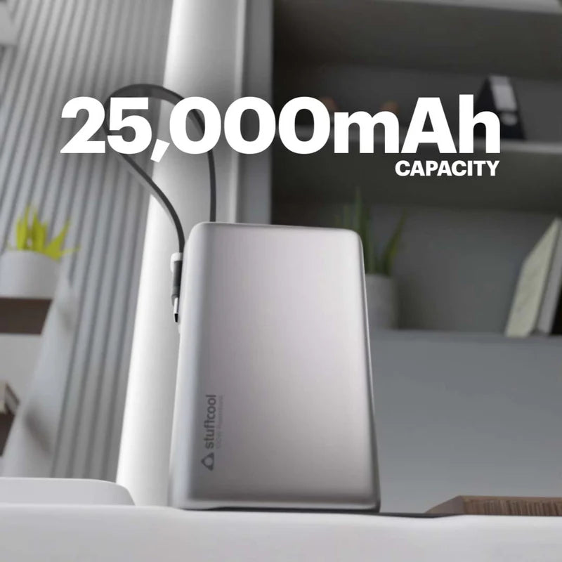 A silver portable power bank sits on a white surface in a modern indoor setting, with large overlaid text reading '25,000mAh CAPACITY' and a small visible 'stuftool' logo on the front.